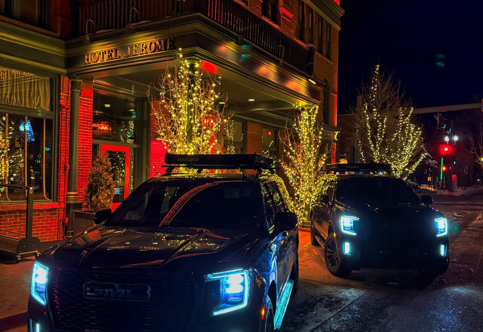 Two black GMC SUVs waiting outside Hotel Jerome in Aspen at night, trees lit with warm lights