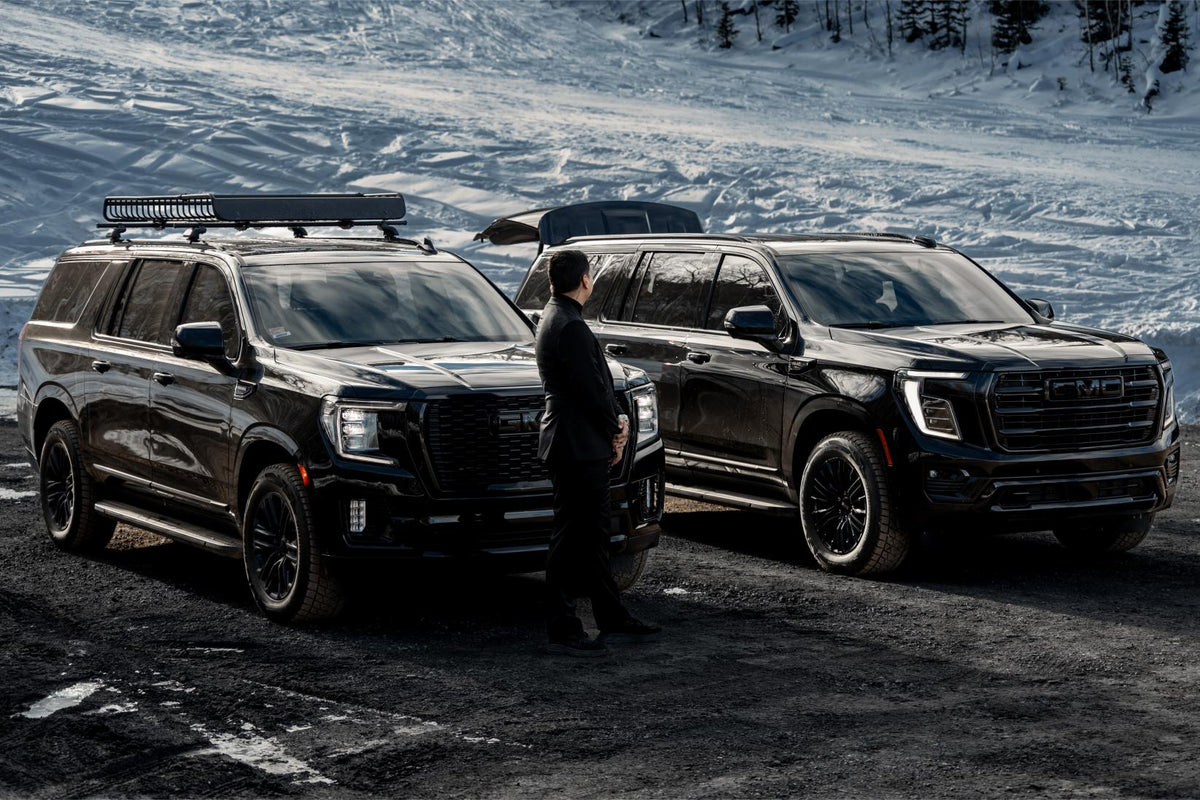 Chauffeur loading luggage into a black GMC Yukon with the tailgate raised, a second SUV alongside on a snowy pullout