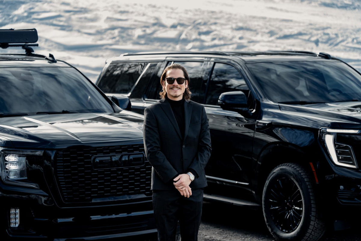 Chauffeur in a black suit and sunglasses standing between two black GMC SUVs on a snowy road