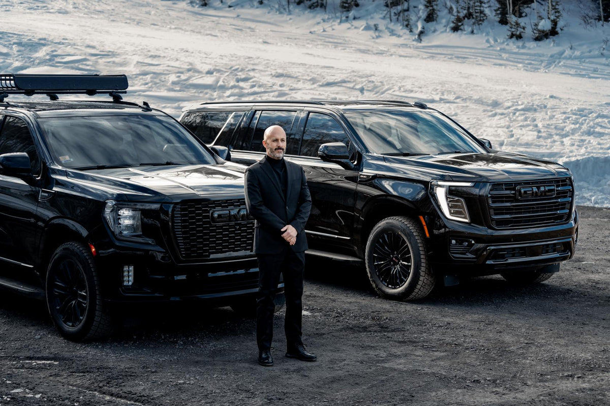 Chauffeur in a black suit standing between two black GMC Yukons parked on packed snow