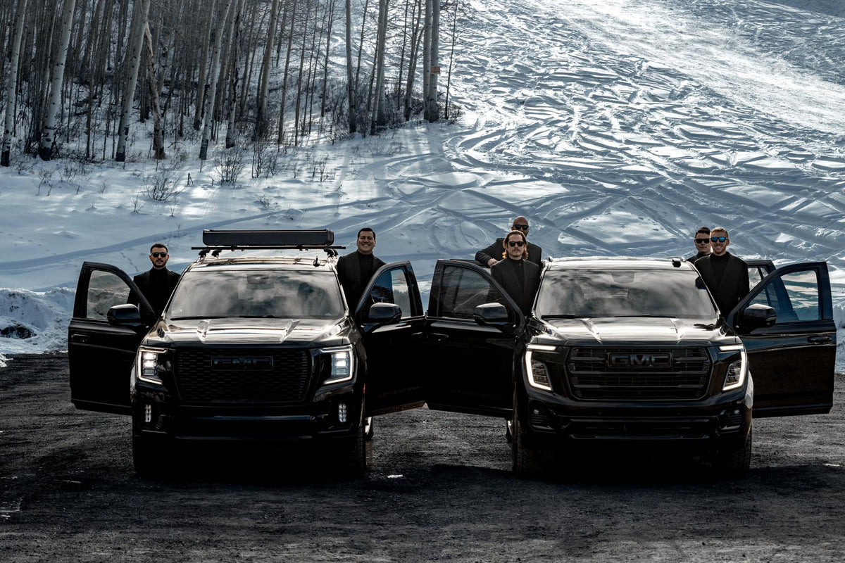 Six chauffeurs standing with the doors open on two black GMC SUVs below a snow-covered ski slope