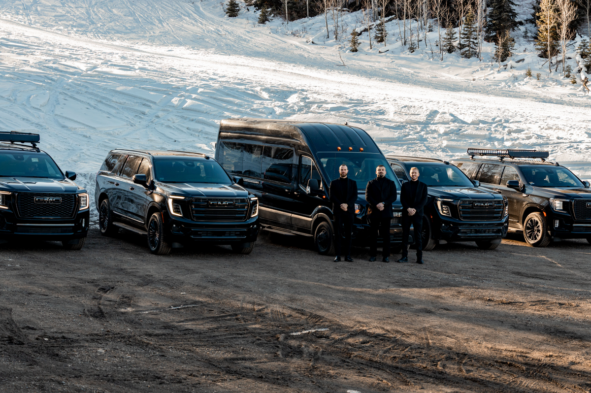 Aspen Empire's fleet of black GMC SUVs and a Ford Transit van lined up on a snow-covered mountain road, three chauffeurs standing in front