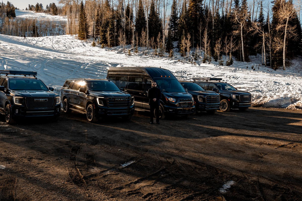 Six black Aspen Empire vehicles parked along a snowy road lined with aspen trees, a chauffeur standing beside them