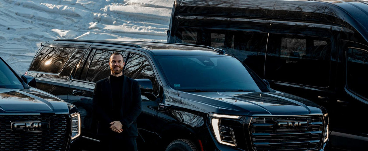 Chauffeur in a black suit standing between two black GMC SUVs on a snow-covered road