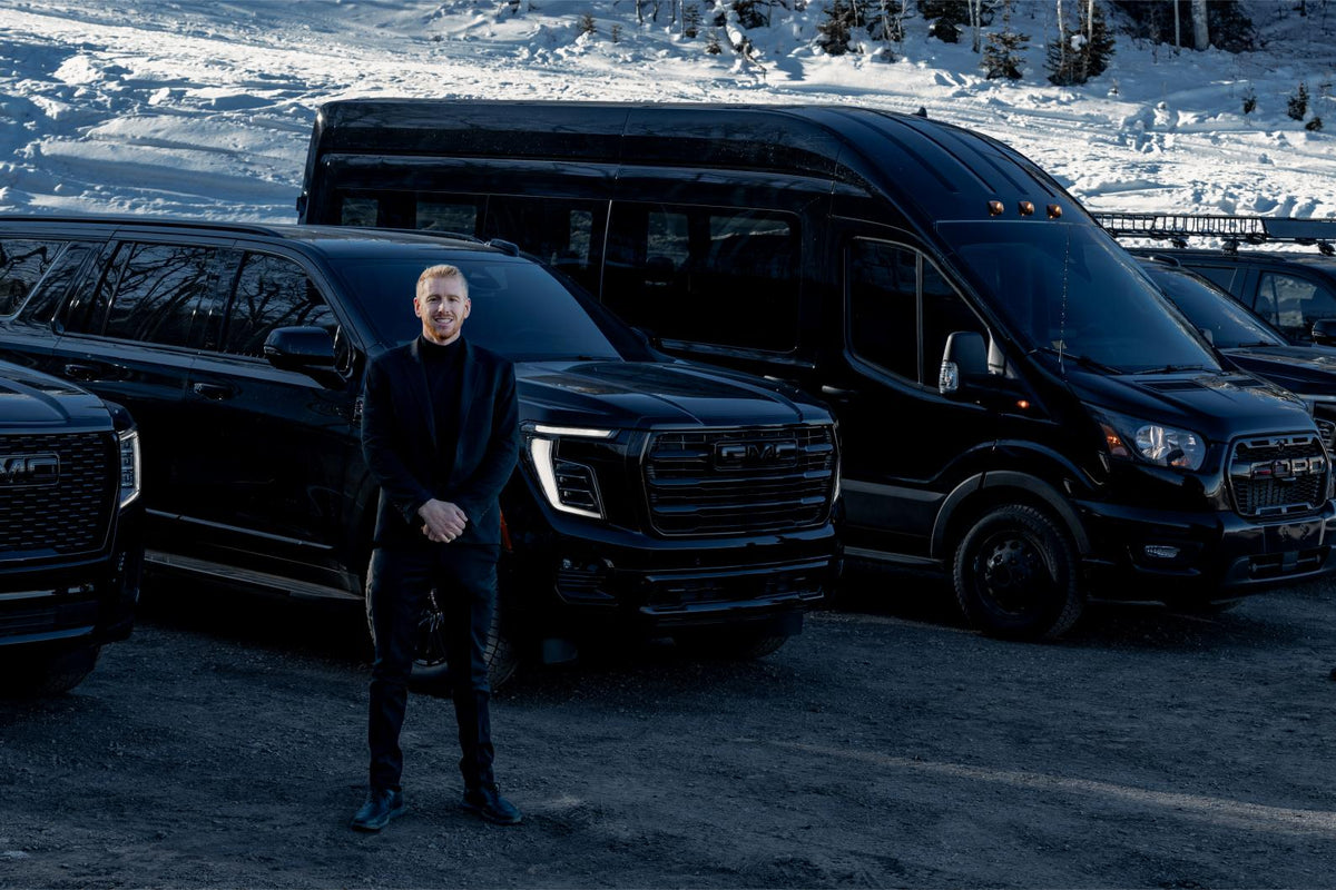 Chauffeur in a black suit standing in front of a black GMC SUV and a Ford Transit van in the snow