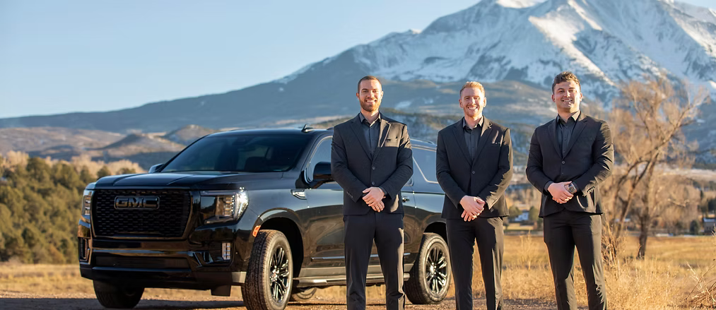 The three founders of Aspen Empire Limousine in black suits beside a black GMC SUV, with Mount Sopris behind them