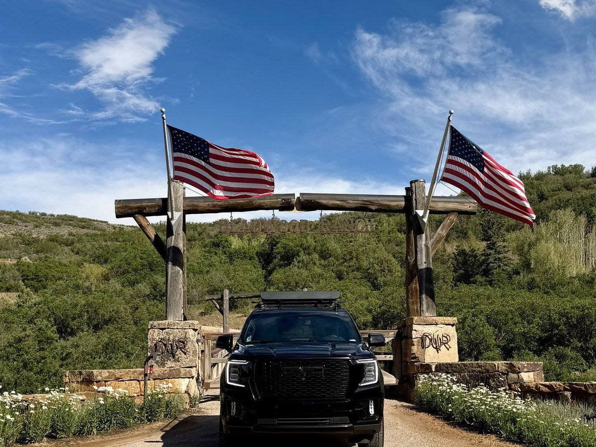 Black GMC Yukon at a log ranch gate flying two American flags in the Colorado high country